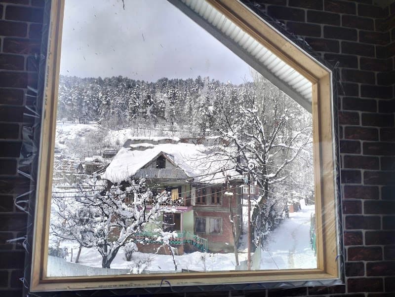 A cozy winter scene framed by a window showcases snow-covered rooftops and trees in a serene mountain village, ideal for travelers seeking a tranquil retreat in Gulmarg.