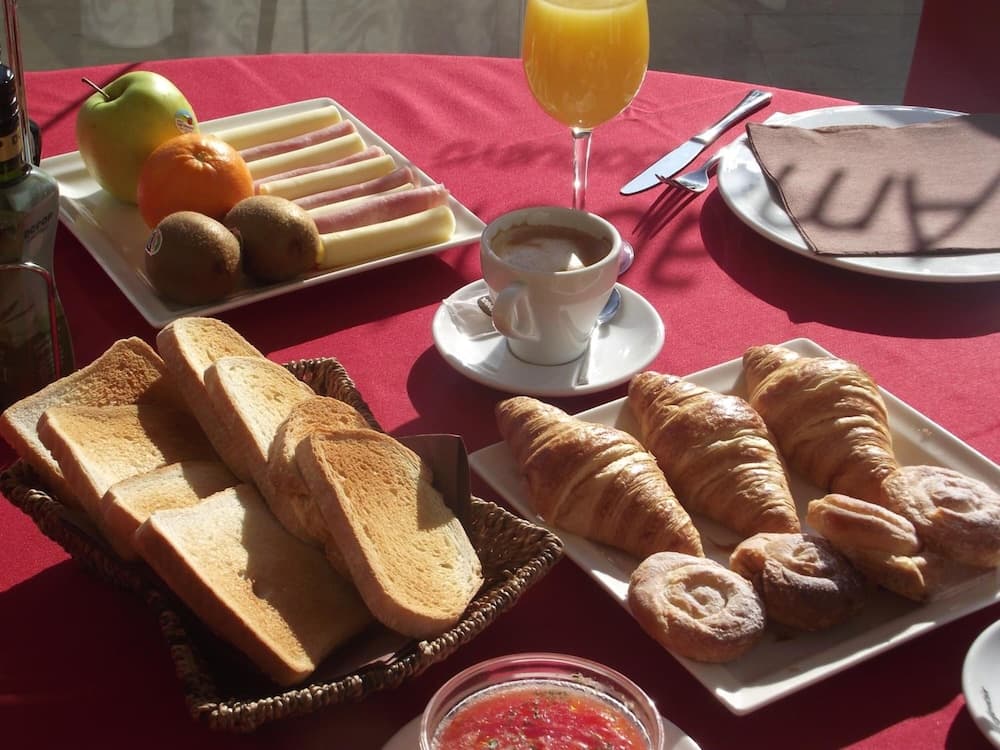 A sunlit breakfast table set with croissants, toast, fresh fruits, coffee, and orange juice on a red cloth creates a cozy morning ambiance ideal for travelers seeking a delightful start to their day.