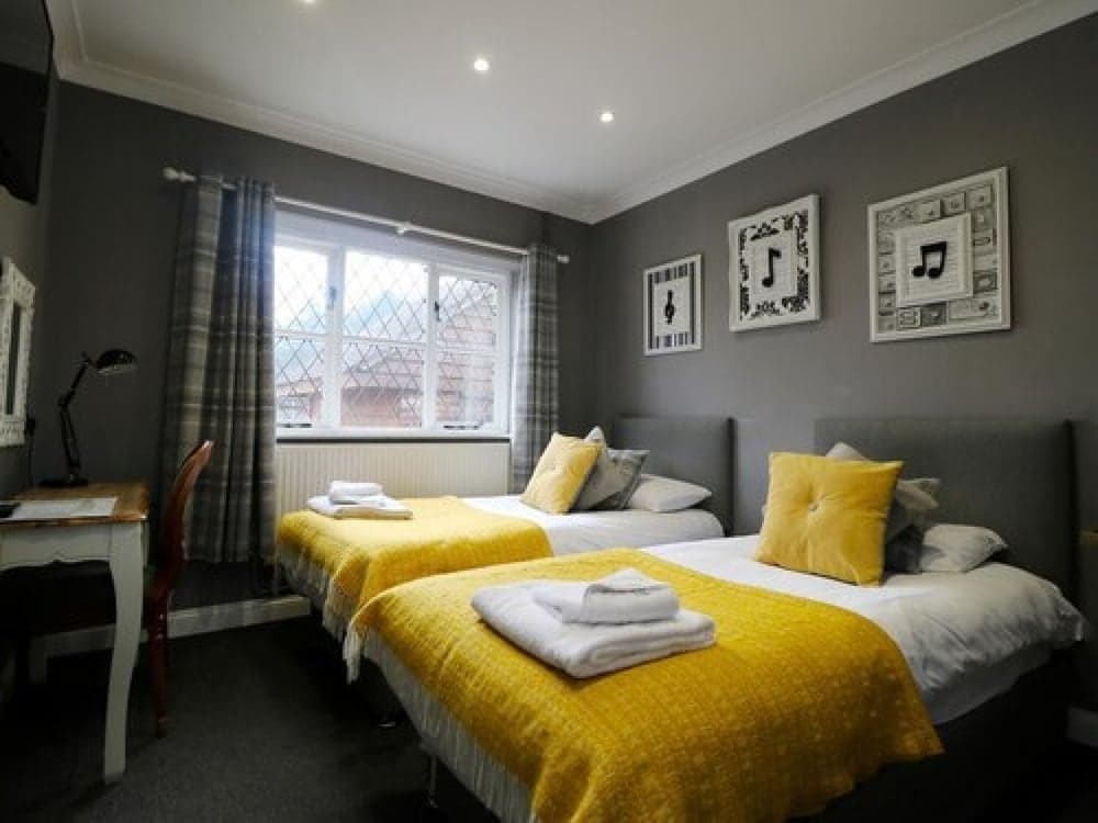 A cozy hotel room features two neatly made beds with yellow throws, gray walls adorned with musical-themed artwork, and a desk by the window, ideal for travelers seeking comfort and charm.