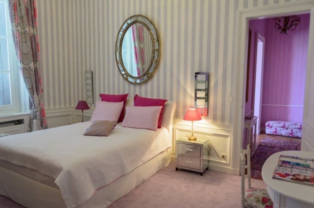 A cozy bedroom with striped wallpaper, a neatly made bed adorned with pink pillows, a round mirror, and a warmly lit bedside lamp creates an inviting atmosphere ideal for a romantic getaway.