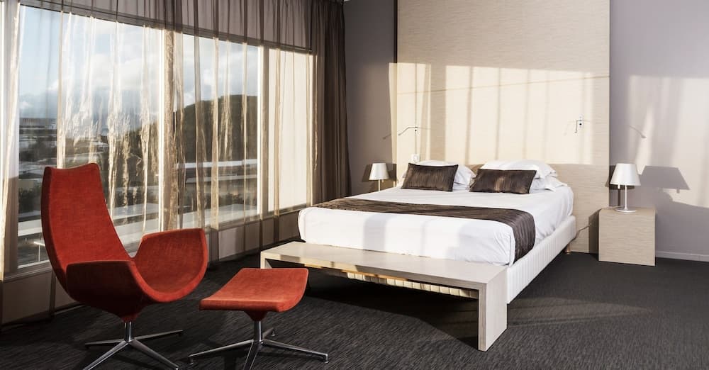 A serene hotel room features a modern bed setup with a vibrant red chair by large windows showcasing a scenic view, ideal for travelers seeking comfort and relaxation.