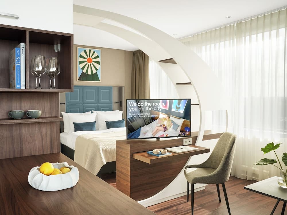 A modern hotel room features a cozy bed, sleek desk setup with a TV displaying city exploration content, and a stylish shelving unit, creating an inviting atmosphere perfect for travelers seeking comfort and convenience.