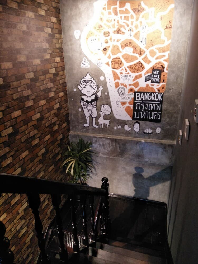A cozy indoor staircase adorned with a brick wall and a vibrant Bangkok-themed mural featuring playful characters and text, ideal for travelers exploring urban art and culture.