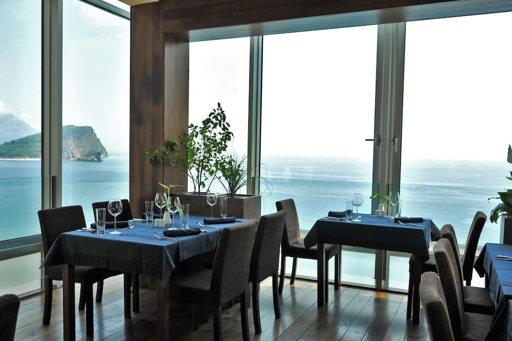 A serene coastal restaurant with large windows framing the ocean view offers an elegant dining experience with neatly set tables and lush greenery, ideal for travelers seeking a luxurious seaside meal.