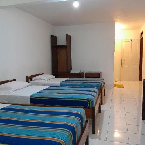 A cozy budget hotel room features three neatly arranged beds with blue striped bedding, a wooden wardrobe, and tiled floors, ideal for travelers seeking simple accommodations in a clean and functional space.