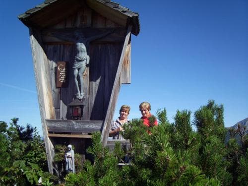 Two travelers pose joyfully beside a rustic wooden chapel adorned with a crucifix, nestled among lush greenery under a clear blue sky, ideal for spiritual retreats or scenic mountain hikes.