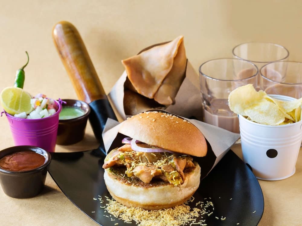 A vibrant street food scene featuring a loaded burger, samosas, potato chips, and colorful condiments on a black plate, perfect for exploring local culinary delights during a travel adventure.