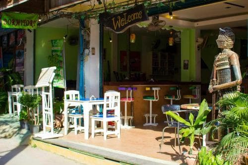 A vibrant outdoor café with colorful stools, white tables, and tropical plants exudes a welcoming and relaxed atmosphere, ideal for travelers seeking a cozy spot to unwind and enjoy local culture.