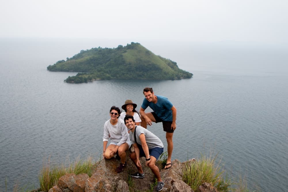 A cheerful group poses on a rocky cliff overlooking a serene lake with a lush green island in the distance, ideal for adventure seekers and nature lovers exploring scenic viewpoints.