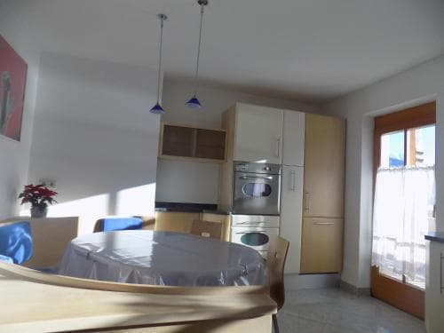 A cozy apartment kitchen with wooden cabinets, a round dining table covered in plastic, and a window letting in natural light, ideal for travelers seeking a homey and functional living space.
