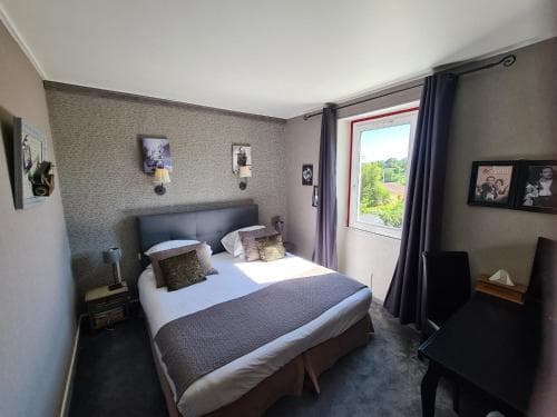 A cozy hotel room with a neatly made bed, framed artwork on textured walls, and a window offering a serene view of greenery, ideal for travelers seeking comfort and relaxation.