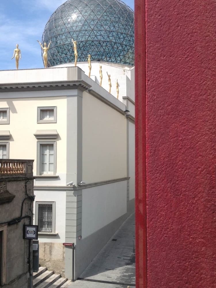 A vibrant urban scene features golden statues atop a white building under a glass dome, framed by a bold red wall, ideal for exploring modern architecture and art in a city setting.