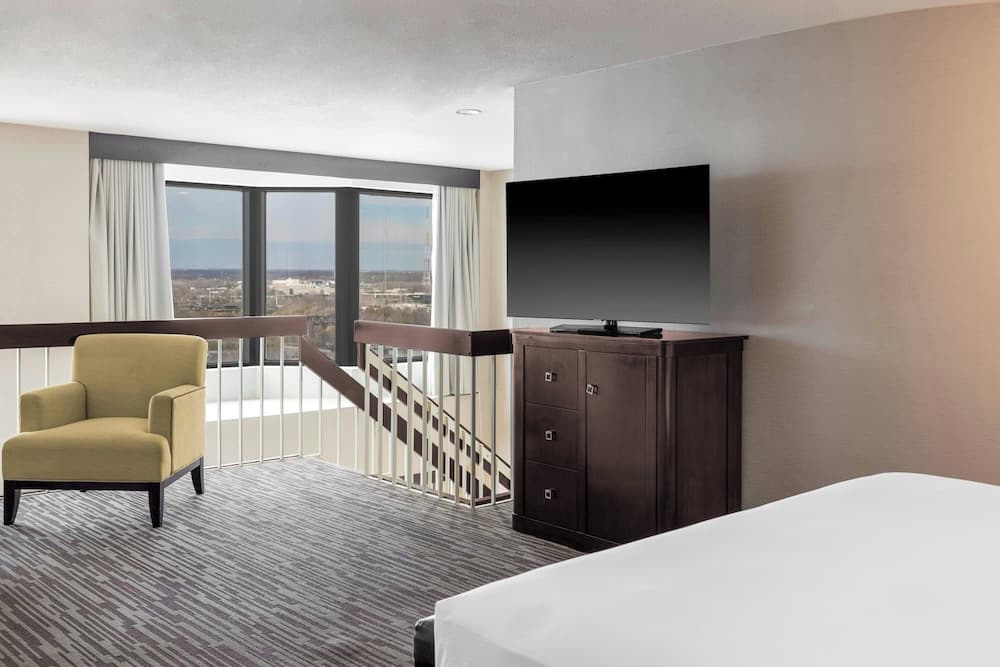 A cozy hotel suite features a comfortable armchair, a sleek TV atop a wooden cabinet, and a panoramic view through large windows, ideal for travelers seeking relaxation and modern amenities.