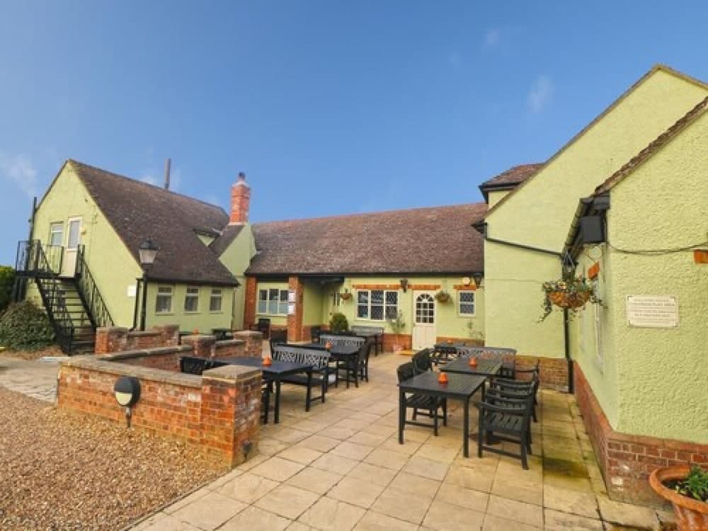 A charming countryside pub with a cozy outdoor seating area, featuring stone-paved grounds, black tables and chairs, hanging flower baskets, and a bright blue sky, perfect for travelers seeking a relaxing dining experience in a quaint setting.