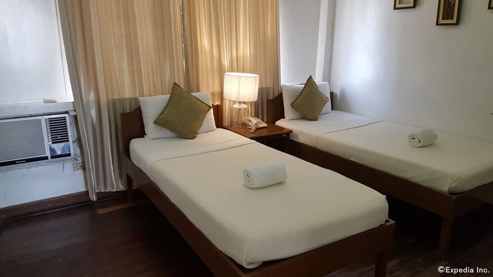 A cozy hotel room features two neatly made single beds with white linens, a soft lamp, and a window with curtains, ideal for budget travelers seeking comfort and simplicity.