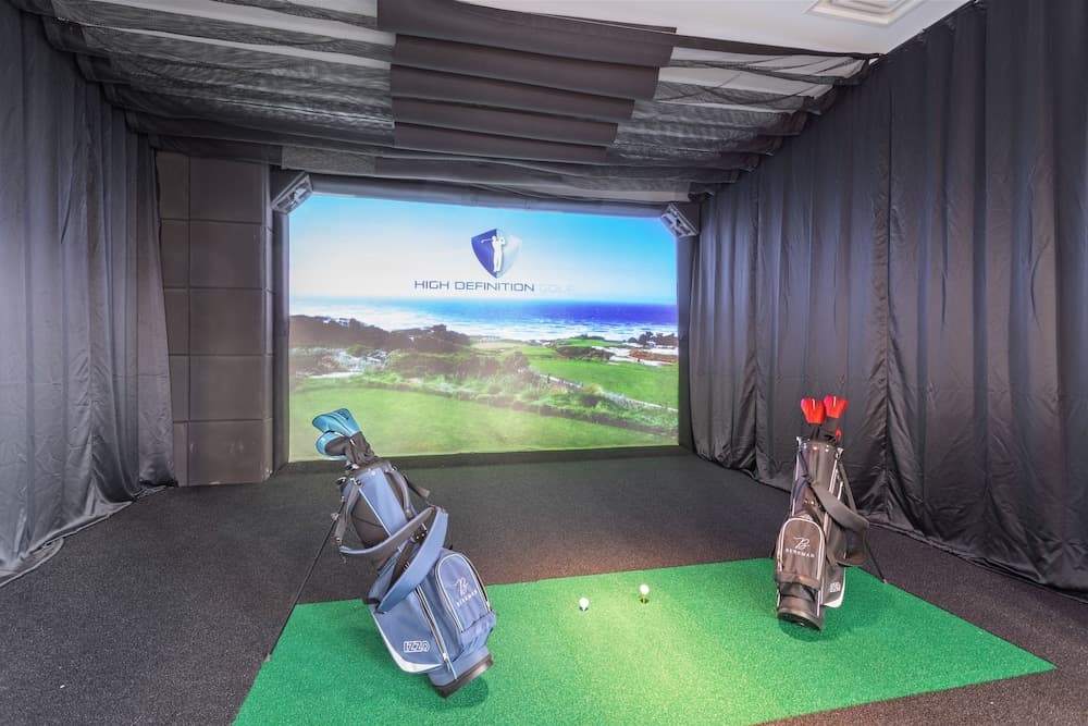 An indoor golf simulator room features a vivid coastal course projection on the screen, surrounded by dark curtains, with two golf bags ready on a green mat, ideal for practicing swings in a controlled environment.