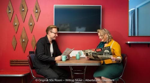 In a vibrant red-walled retro diner, two guests enjoy coffee and magazines at a cozy table, evoking a nostalgic 50s ambiance perfect for a themed getaway or vintage-inspired travel experience.