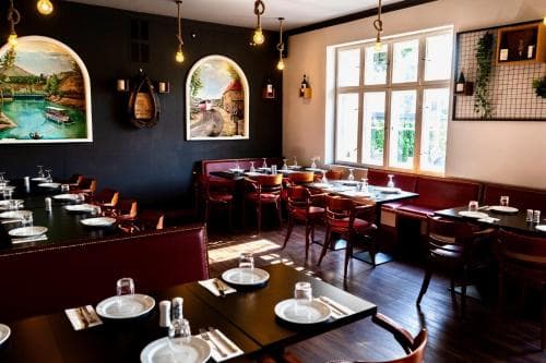 A cozy restaurant interior features rustic wooden tables set for dining, warm pendant lighting, and charming wall murals, creating an inviting atmosphere perfect for travelers seeking a relaxing meal experience.