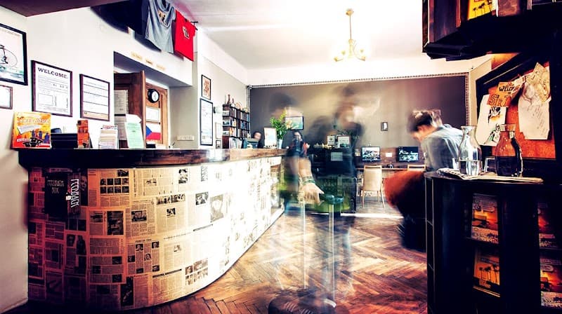 A cozy hostel reception in Prague features a warmly lit bar adorned with newspaper clippings, creating an inviting atmosphere for travelers seeking local charm and comfort.