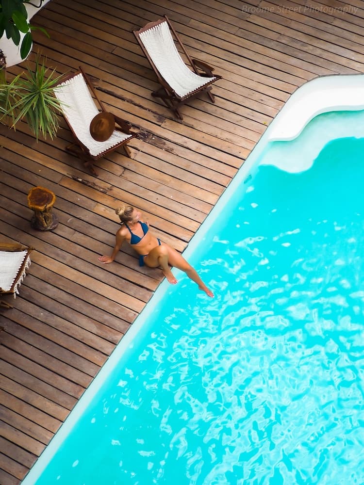 A serene poolside scene features a woman relaxing by the edge of a turquoise pool on a wooden deck adorned with lounge chairs and potted plants, ideal for promoting luxury vacation retreats.