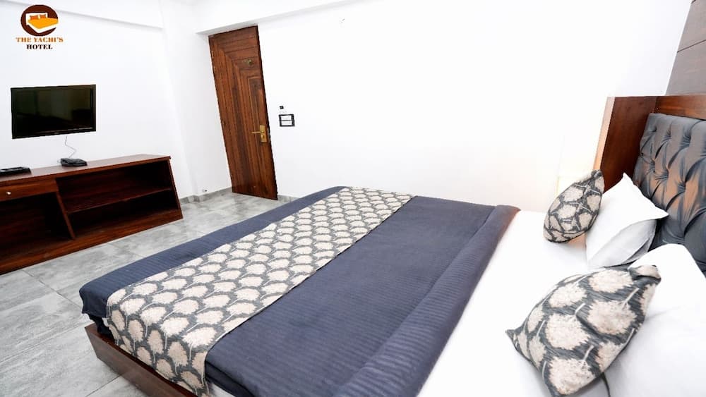 A cozy hotel room features a neatly made bed with patterned pillows, a wooden door, a mounted TV on a console, and tiled flooring, ideal for travelers seeking comfort and simplicity.