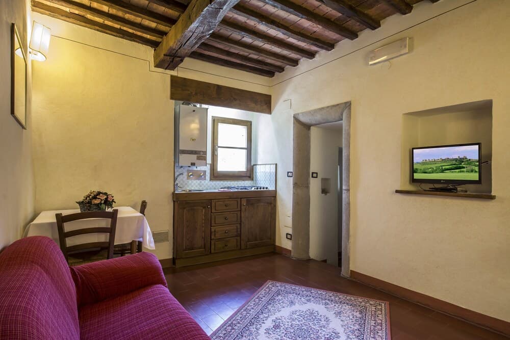 A cozy rustic apartment features exposed wooden beams, a small kitchenette, a red sofa, and a flat-screen TV, ideal for travelers seeking a charming and comfortable stay in a historic setting.