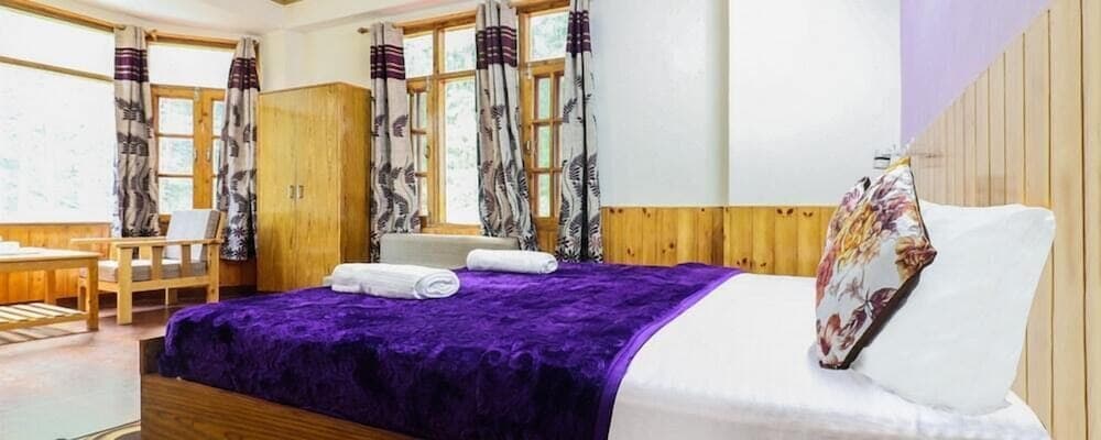 A cozy bedroom with wooden furnishings, purple accents, and large windows offering a serene view, ideal for travelers seeking relaxation in a rustic retreat.