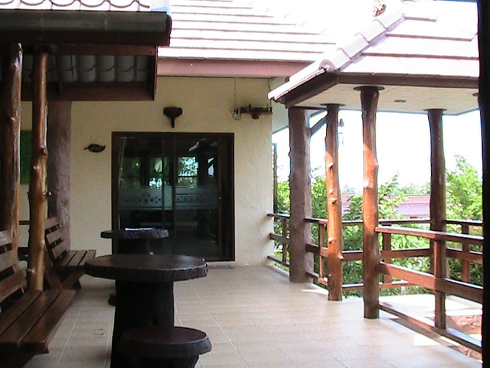 A serene outdoor patio features rustic wooden furniture under a tiled roof, offering a tranquil retreat for travelers seeking relaxation amidst lush greenery.