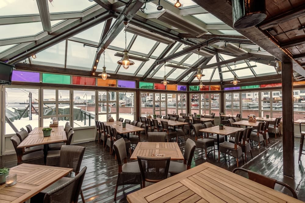 A cozy waterfront restaurant with wooden tables, colorful glass panels, and panoramic views offers a serene dining experience ideal for travelers seeking relaxation and scenic ambiance.