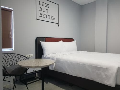 A minimalist hotel room features a neatly made bed beside a small table and chair, with a motivational wall sign reading "LESS BUT BETTER," ideal for travelers seeking a cozy and modern retreat.
