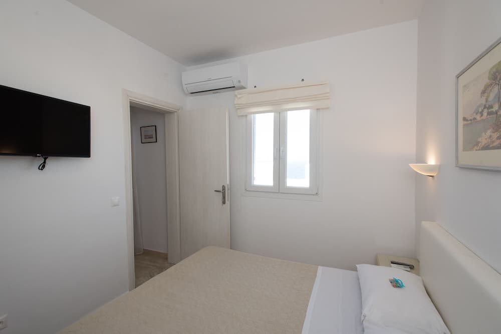 A serene hotel bedroom with minimalist decor, featuring a neatly made bed, wall-mounted TV, air conditioning unit, and a window with a view, ideal for travelers seeking comfort and simplicity.