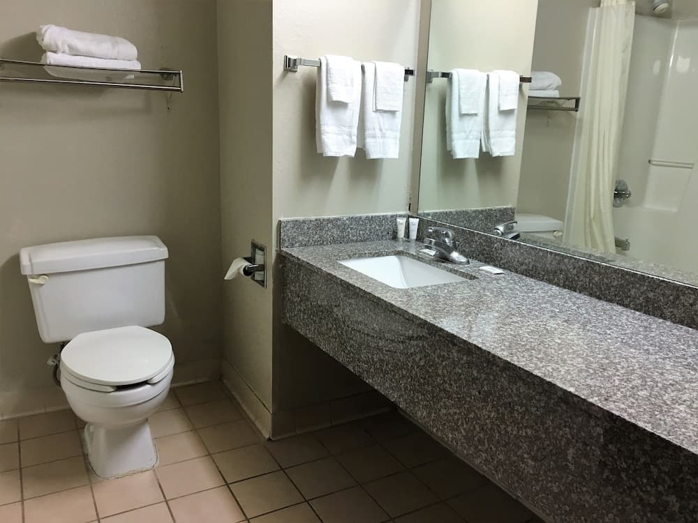 A clean hotel bathroom features a tiled floor, granite countertop, white fixtures, neatly folded towels, and a large mirror, ideal for travelers seeking comfort and convenience.