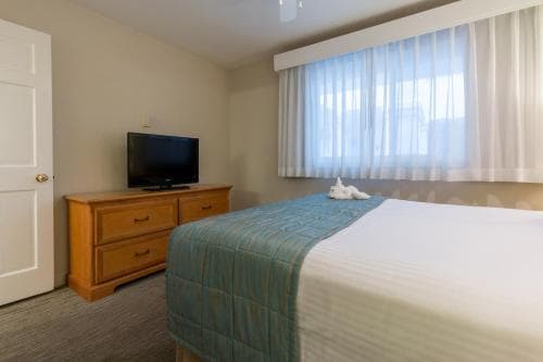 A cozy hotel room features a neatly made bed with a blue blanket, a wooden dresser topped with a TV, sheer curtains over a window, and a welcoming atmosphere ideal for travelers seeking comfort and relaxation.