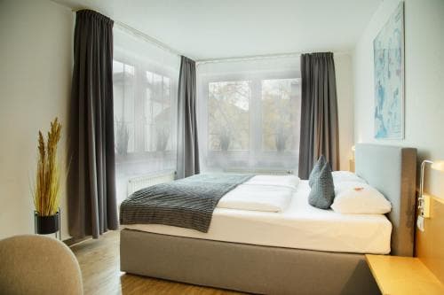 A cozy hotel room with a neatly made bed, large windows offering a serene view, modern decor including gray curtains and artwork, ideal for travelers seeking comfort and relaxation.