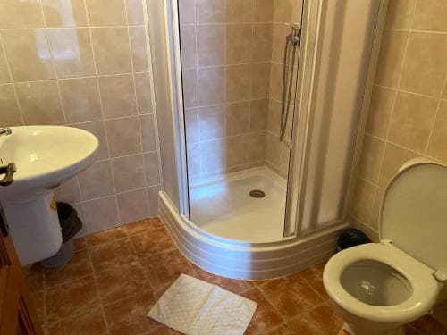 A cozy bathroom with beige tiled walls, a corner shower, white sink, toilet, and a bath mat creates a clean and functional space ideal for travelers seeking comfort in accommodations.