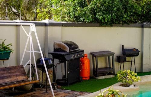 A cozy backyard patio features a barbecue setup with a grill, propane tank, and fire pit beside a pool, surrounded by lush greenery, ideal for outdoor gatherings and relaxation.