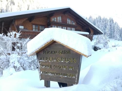 A cozy snow-covered chalet nestled in a serene winter landscape, featuring a charming signboard for Pension Garni Alpengruss, ideal for travelers seeking a peaceful mountain retreat.