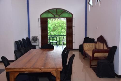 A cozy indoor dining area with a wooden table, black chairs, and a wicker sofa set against white walls, features an arched window offering a lush green view, ideal for travelers seeking a tranquil retreat or a quiet meeting space.