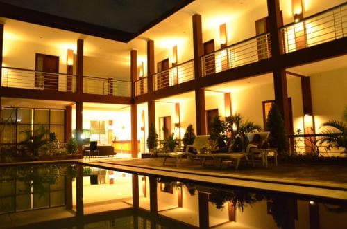 A serene evening view of a well-lit hotel courtyard featuring a reflective pool, lounge chairs, and tropical plants, ideal for travelers seeking relaxation and comfort.