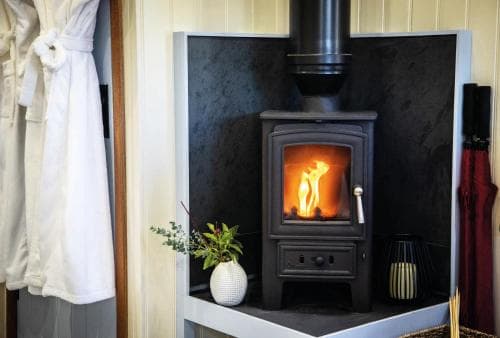 A cozy corner featuring a glowing wood-burning stove set against a dark backdrop, complemented by a white vase with greenery and bathrobes nearby, evokes warmth and relaxation ideal for a boutique hotel retreat.