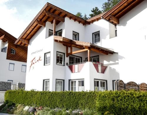 A charming white apartment building with wooden accents and balconies nestled among lush greenery offers a cozy retreat in an alpine setting, ideal for travelers seeking relaxation and scenic views.