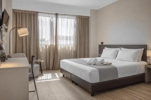 A cozy hotel room with a neatly made bed, soft lighting, and large windows draped in beige curtains creates a serene atmosphere, ideal for travelers seeking relaxation and comfort.