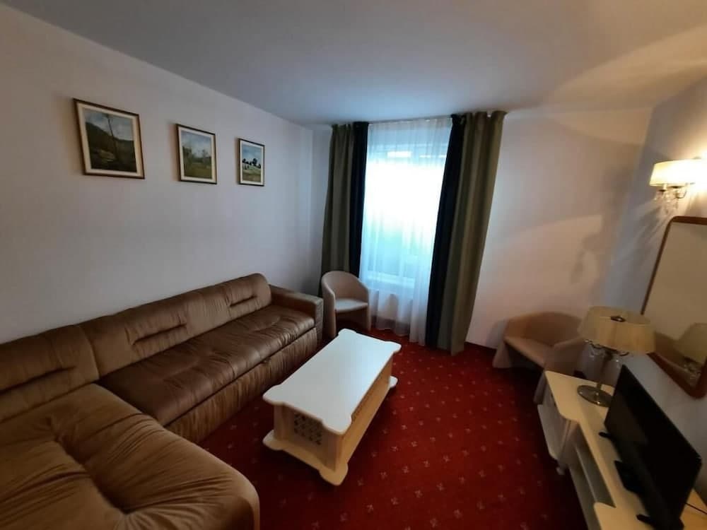 A cozy hotel room features a brown leather sofa, red patterned carpet, framed wall art, and a bright window with curtains, creating a warm and inviting atmosphere ideal for travelers seeking comfort.