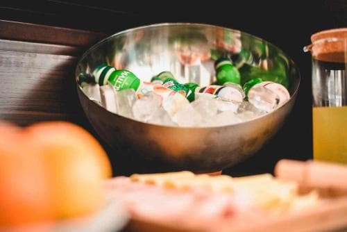 A refreshing scene of a metallic bowl filled with ice and green bottles, set against a cozy indoor backdrop, evokes a relaxed gathering mood ideal for sharing drinks and snacks during a casual get-together.