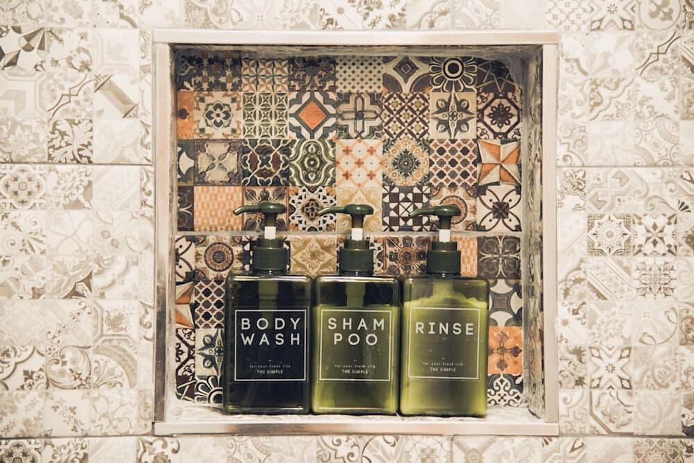 A cozy bathroom niche adorned with intricate patterned tiles displays three sleek bottles labeled "BODY WASH," "SHAMPOO," and "RINSE," perfect for travelers seeking stylish and functional amenities in boutique accommodations.
