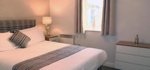A cozy hotel room features a neatly made bed with plaid accents, soft lighting from a bedside lamp, and a window with curtains, creating a welcoming atmosphere ideal for travelers seeking comfort and relaxation.