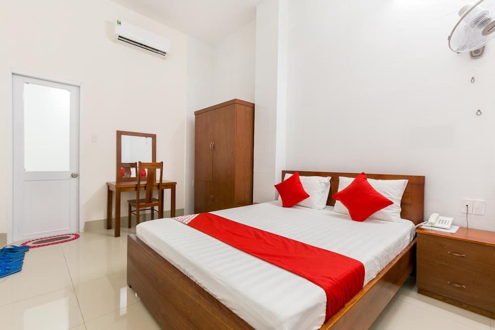 A cozy hotel room features a neatly made bed with red accents, wooden furniture, and a clean white interior, ideal for travelers seeking comfort and simplicity.