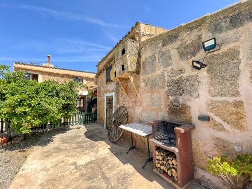 A serene courtyard with rustic stone walls, a barbecue grill stocked with wood, and lush greenery under a clear blue sky offers an idyllic setting for a relaxing vacation retreat.