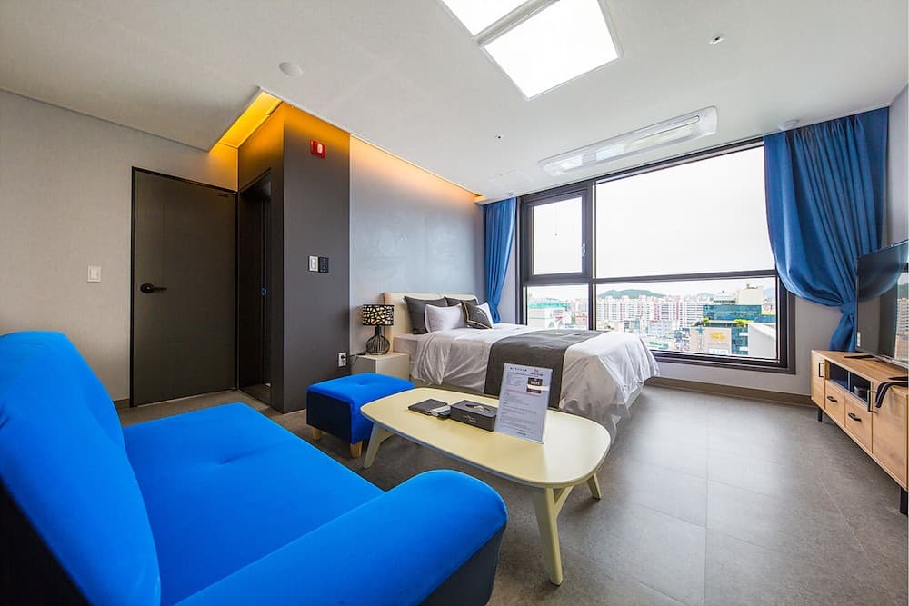 A modern hotel room with a vibrant blue sofa, sleek furnishings, and a panoramic city view through large windows offers a cozy yet stylish retreat for travelers seeking comfort and urban scenery.