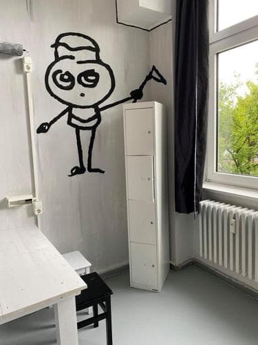 A minimalist hostel room features a quirky wall mural of a cartoon character, a white storage cabinet, and a simple desk by the window, creating a cozy and artistic atmosphere ideal for budget travelers seeking unique accommodations.